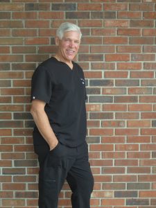 Profile Image of Board-Certified Plastic Surgeon Dr. Paul M. Parker, leaning against a brick wall in black scrubs.